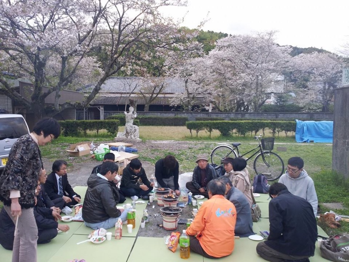 桜並木の下で地区の人たちが集まって花見の食事会をしている様子