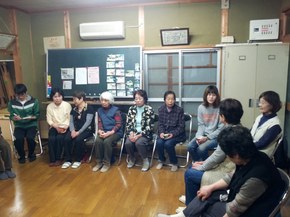 集会所で地区の人たちが輪になって座り、お茶を飲みながら話し合っている様子